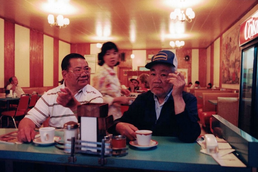 Photo: Noisy Jim wearing baseball cap and smoking a cigarette, at counter in his restaurant, drinking coffee with a friend