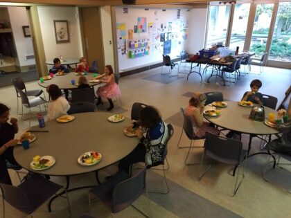children seated around tables in a common hall eating dinner. A table full of craft materials sits at the back.